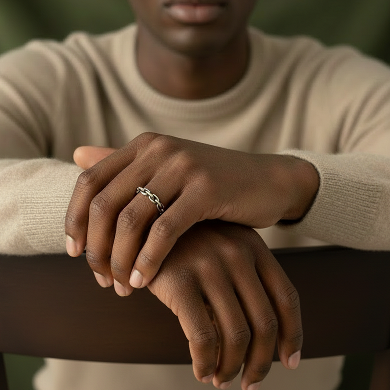 dark skinned model wearing a bege sweater is sitting on a chair and showing his hands and he is wearing a silver chain ring